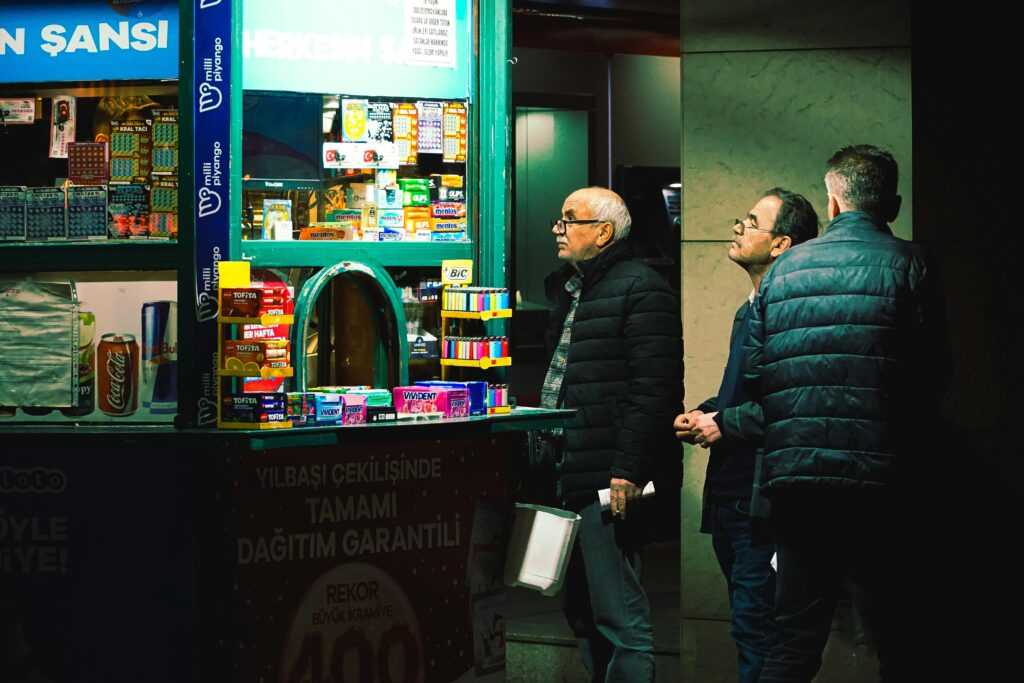Three men at a lottery stand observe the variety of items for sale under bright lights.