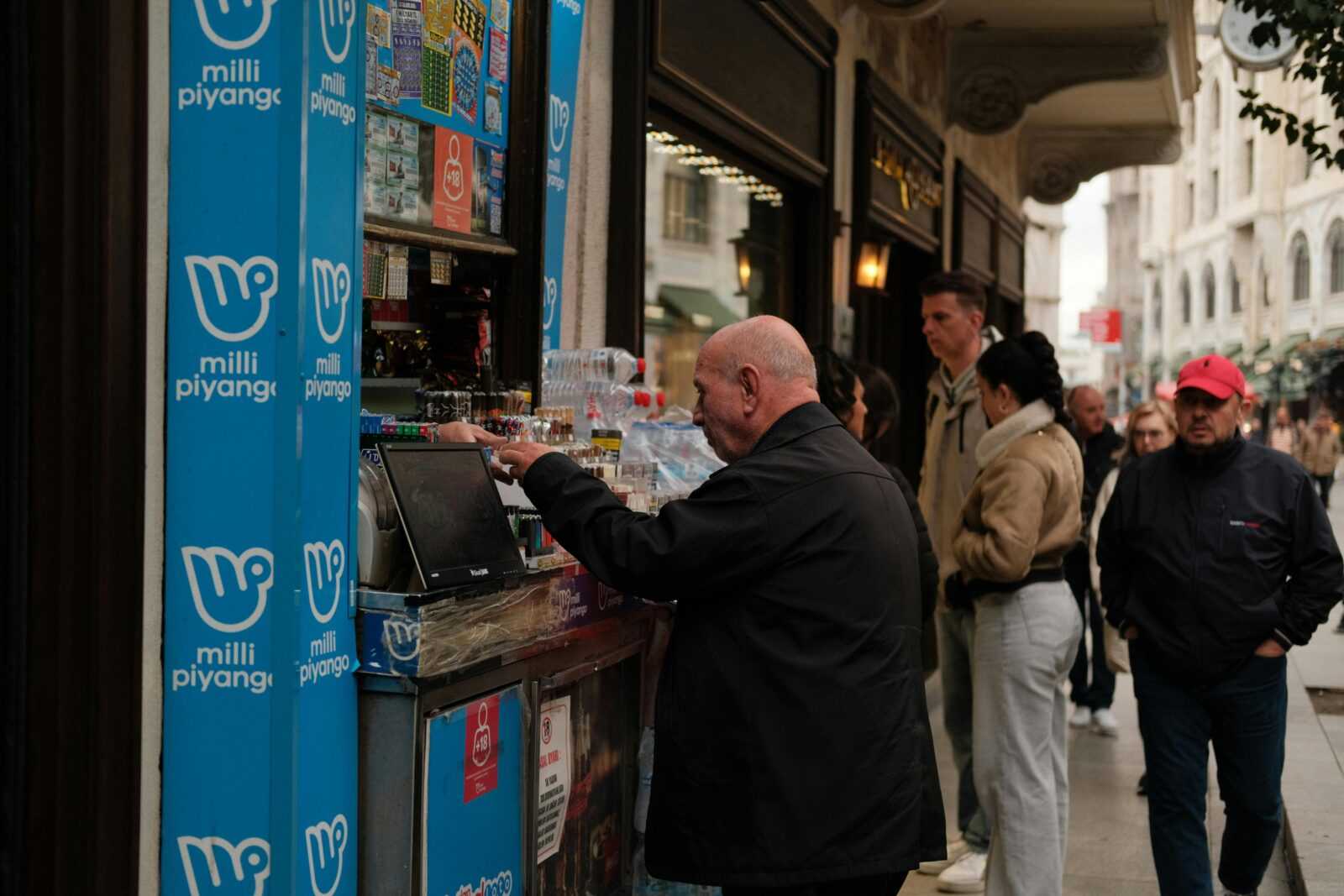 People buying lottery tickets and beverages from a street vendor on a bustling urban street.